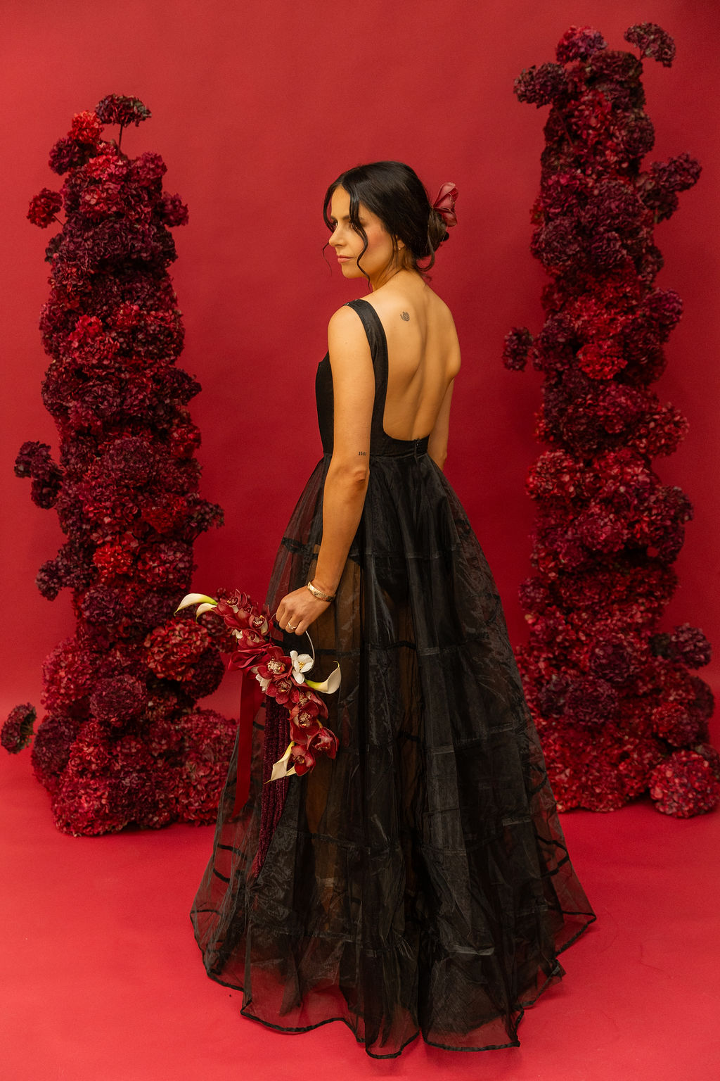 Model in a black tulle gown with an open back between Alanah Conner's dried red floral arches, photographed by Ellen Cayford