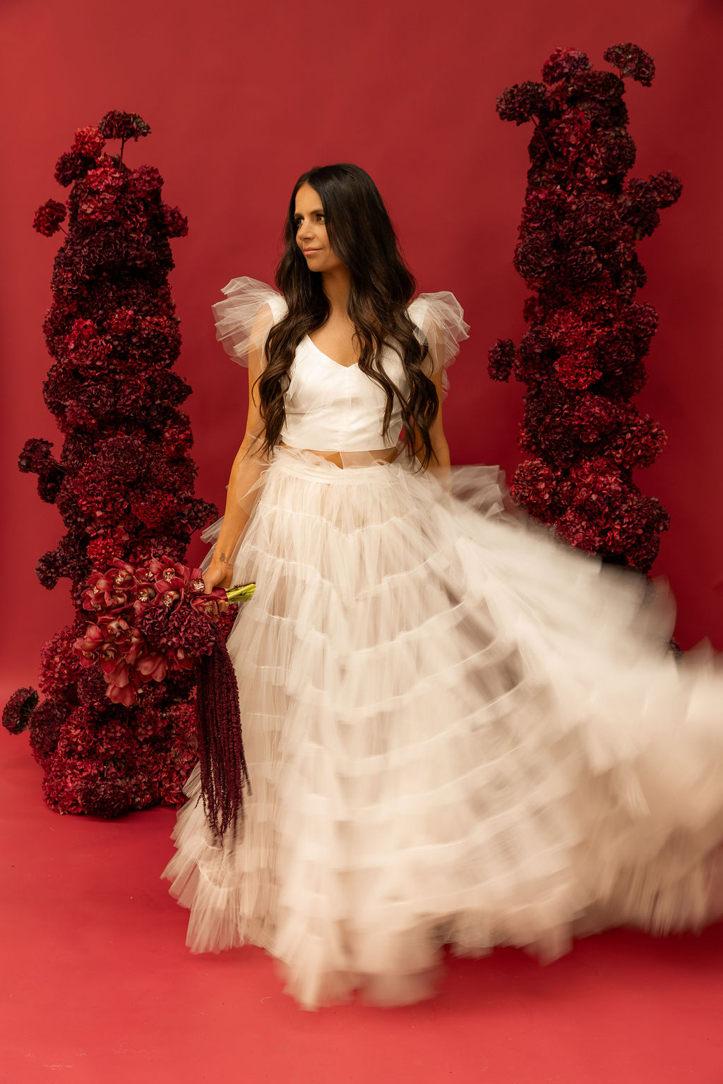 Model spinning in a tulle two-piece against the red backdrop, tulle skirt caught mid-motion, photographed by Ellen Cayford