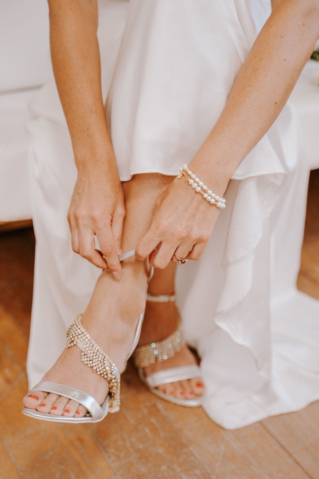 Bride fastening a jewelled bridal sandal beneath her gown — the shoes the hem is set from, needed at every fitting